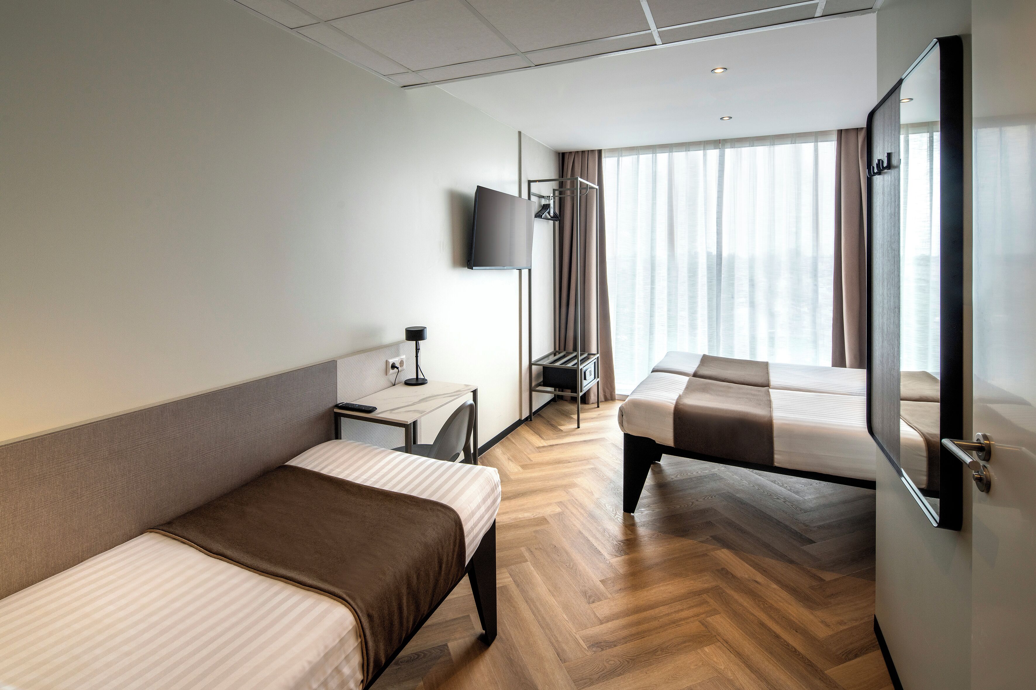 Ett hotellrum med två sängar, ett skrivbord, en stol, en tv som är monterad på väggen och ett stort fönster med gardiner.