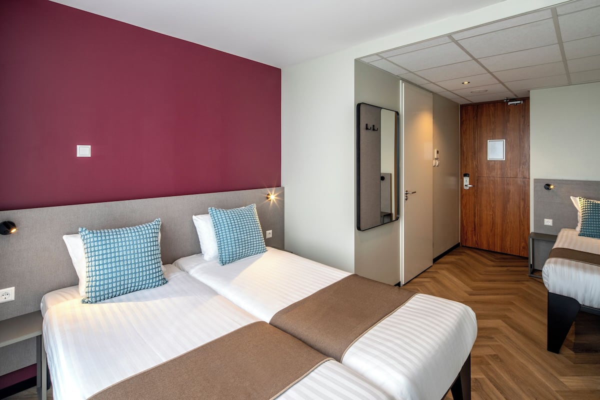 A hotel room with two beds, a wooden floor, a red accent wall, and a mirror.