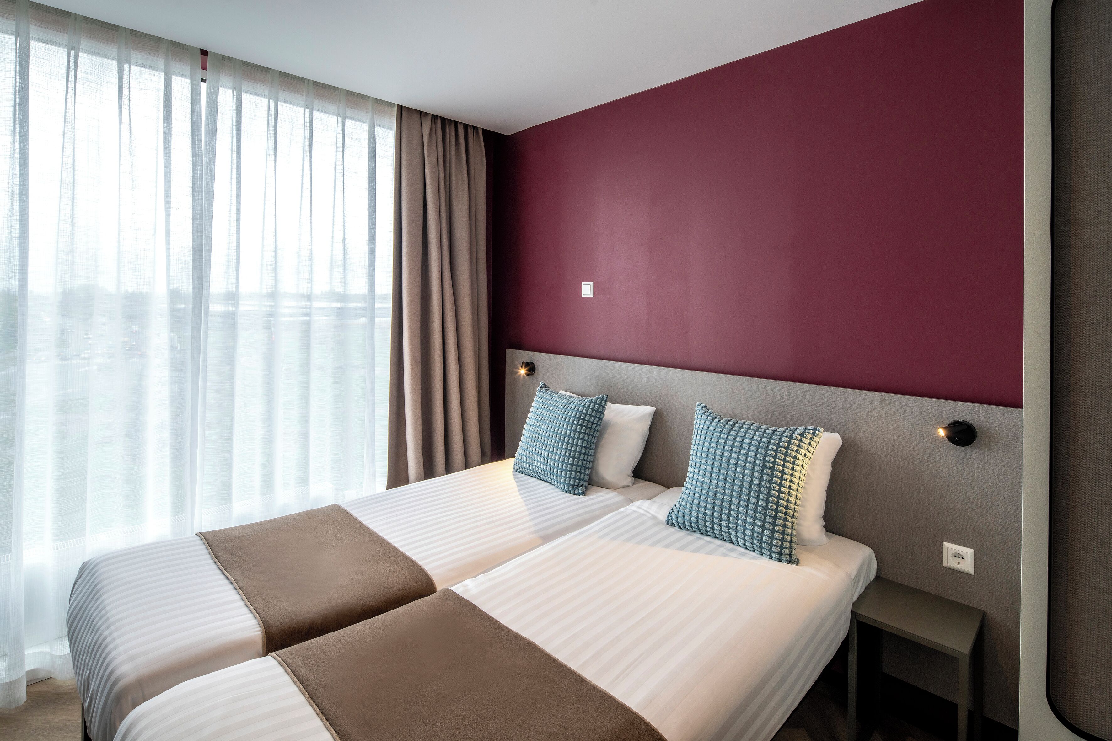 A hotel room with two beds, a red accent wall, and a view through sheer curtains.