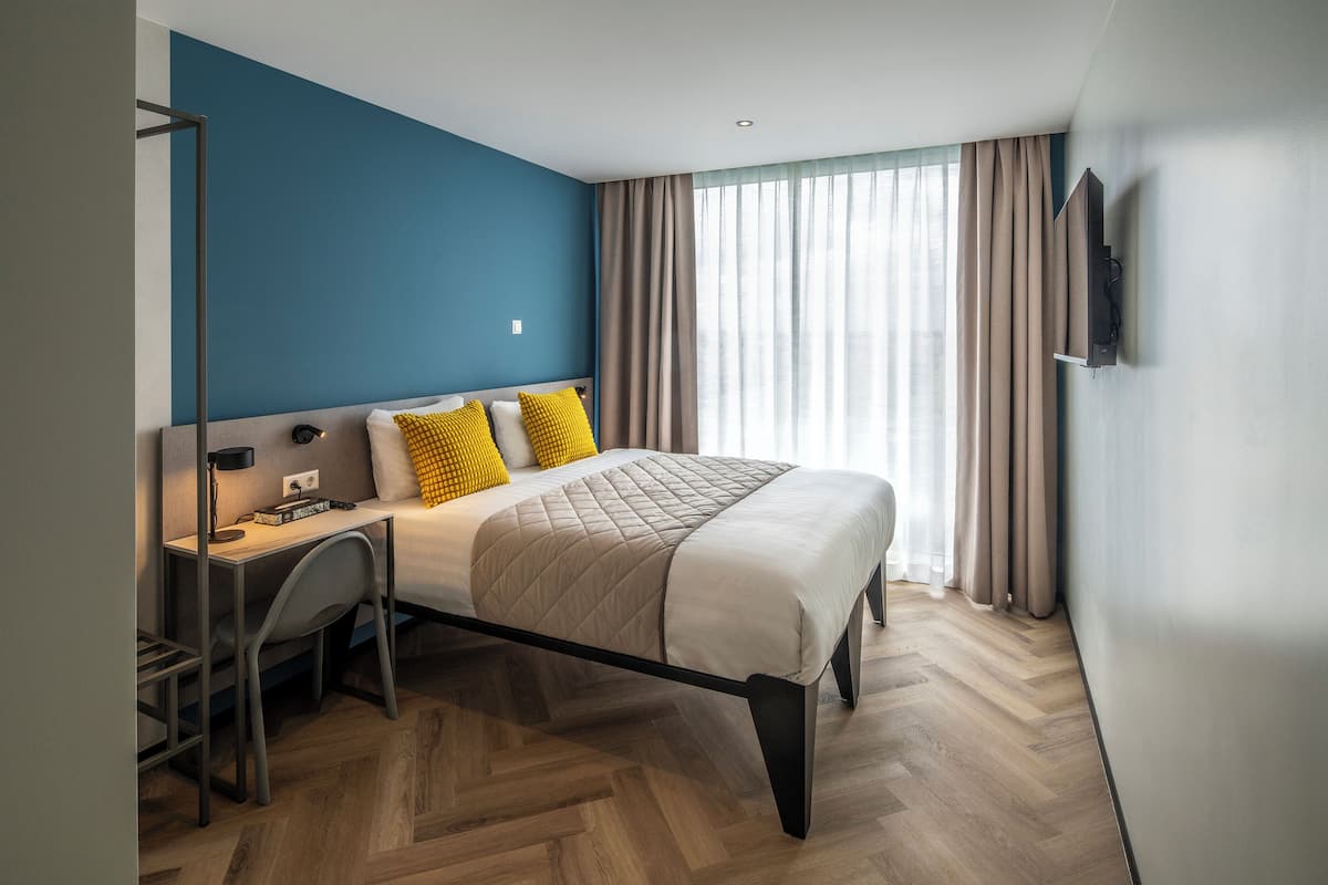 A modern hotel room with a bed, desk, and chair, a television mounted on the wall, and a large window with curtains.
