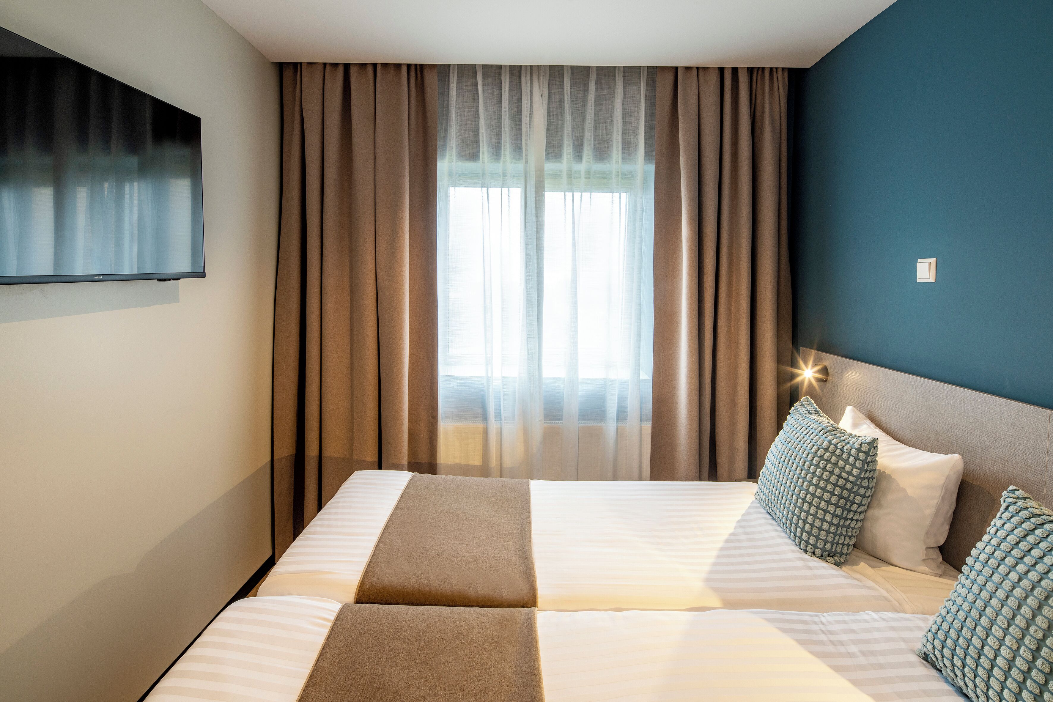 A hotel room with a large bed, a flat-screen TV mounted on the wall, and a window with curtains.