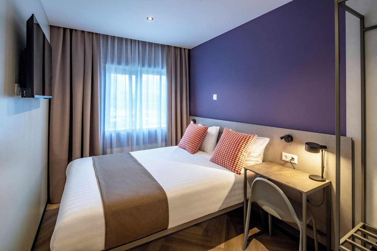 A modern hotel room with a large bed, a desk with a chair, a TV mounted on the wall, and a window with curtains.