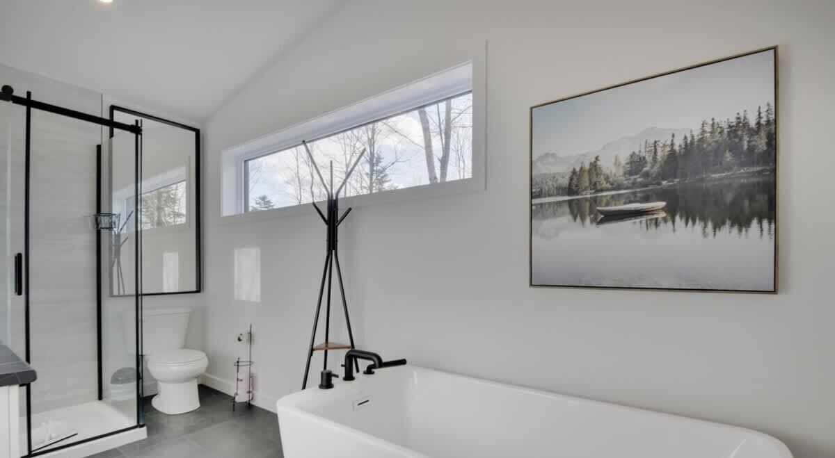 Une salle de bain moderne avec une douche à l’italienne en verre, une baignoire blanche et une grande fenêtre donnant sur un lac et des montagnes.