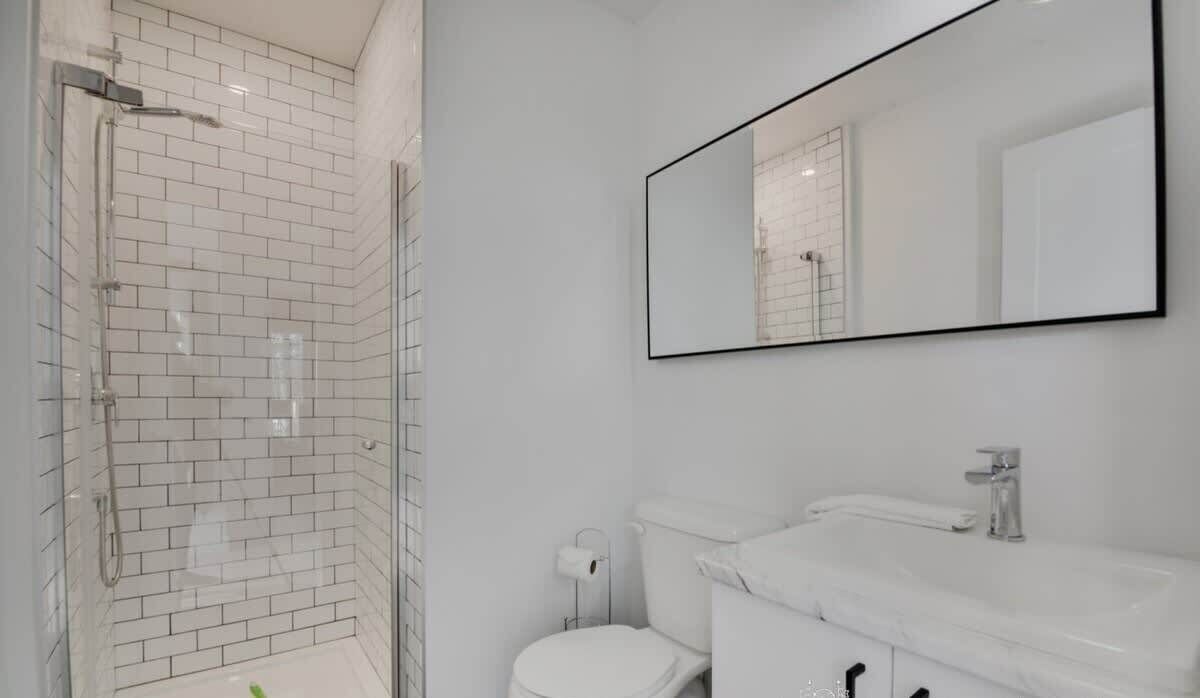 A modern bathroom with white tiles, a glass-enclosed shower, a toilet, and a large mirror.