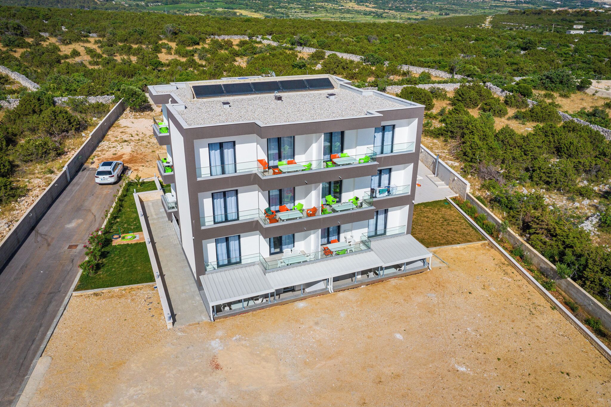 A modern multi-story building with balconies, solar panels, and a view of a green landscape.