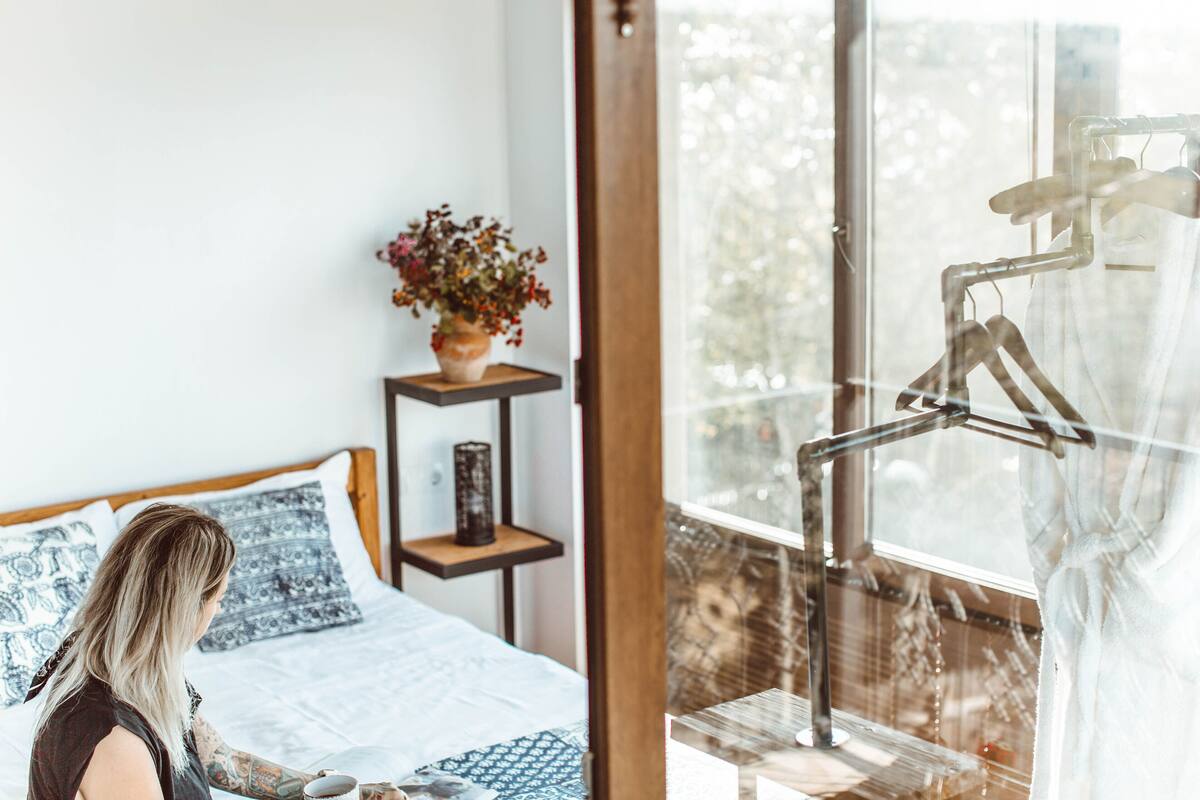 Une chambre à coucher avec un lit, une table de chevet ornée d’un vase de fleurs et une fenêtre avec un rideau.