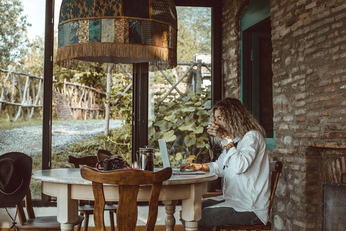 Une personne travaille sur un ordinateur portable, assise à une table ronde en bois, sous une lampe au style rustique.