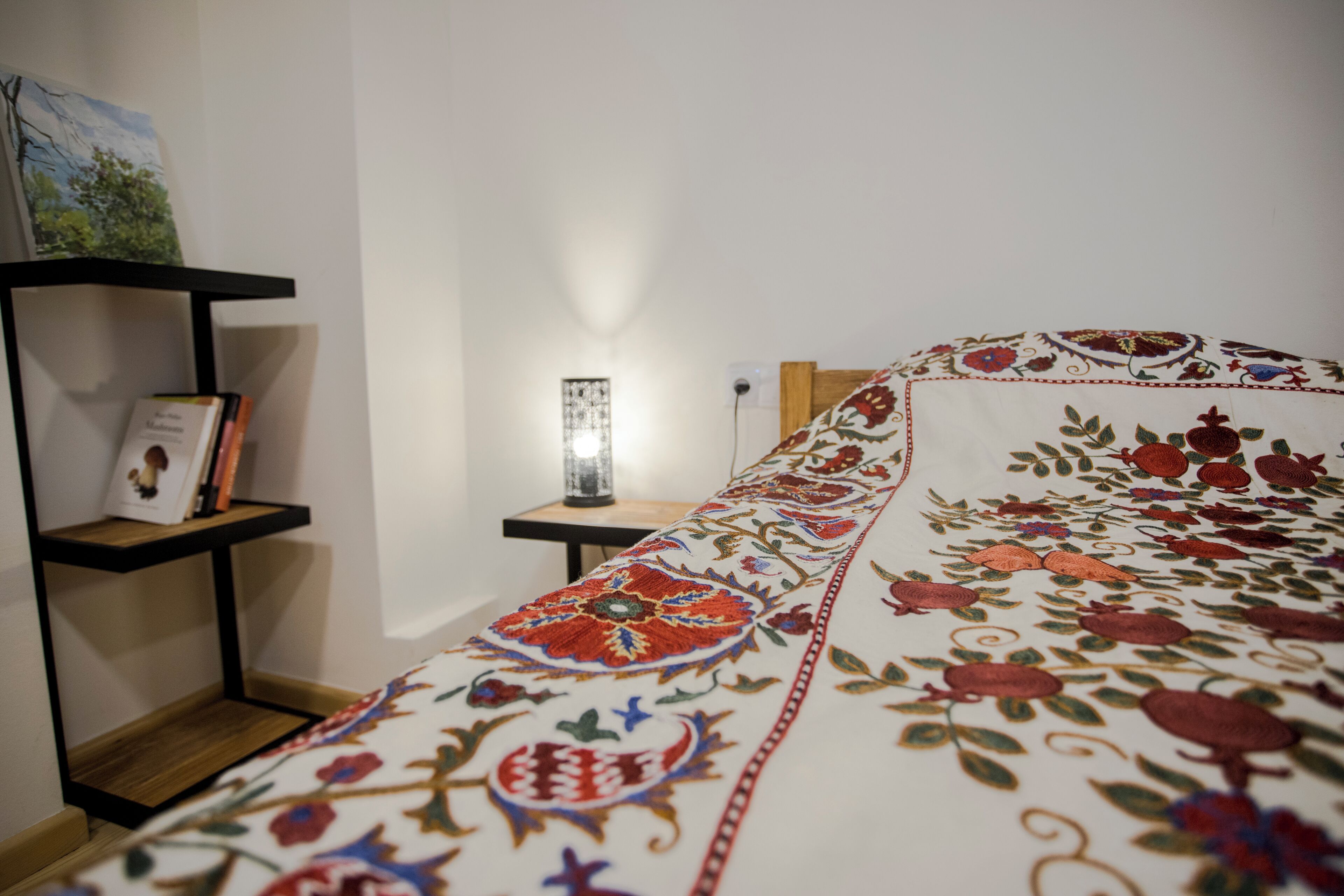 A neatly made bed with a floral-patterned bedspread, a bedside table with a lamp, and a bookshelf against the wall.