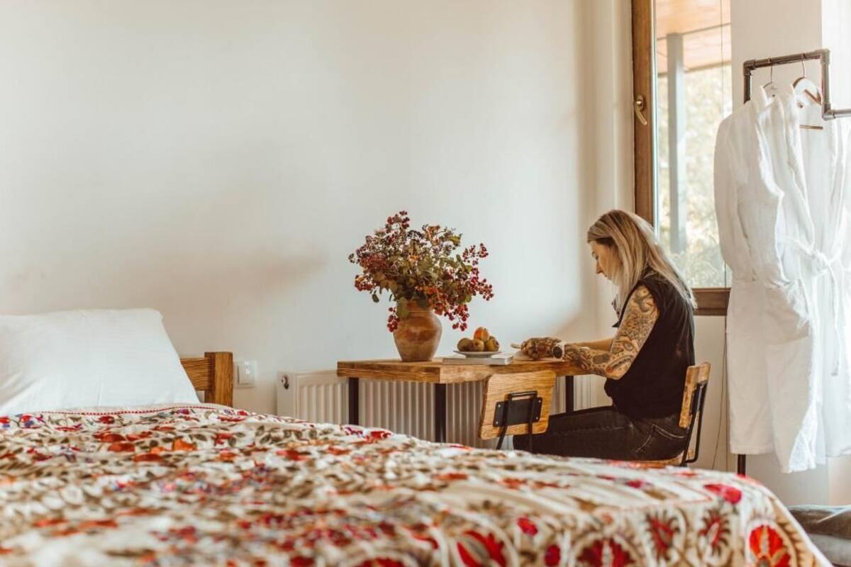 Une pièce comprenant un lit, une table en bois ornée d’un vase de fleurs et une fenêtre à laquelle est accrochée une robe blanche.