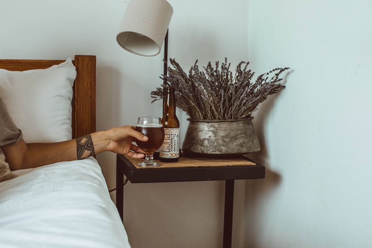 Une personne assise sur un lit, avec une bouteille et un verre posés sur une table de chevet.