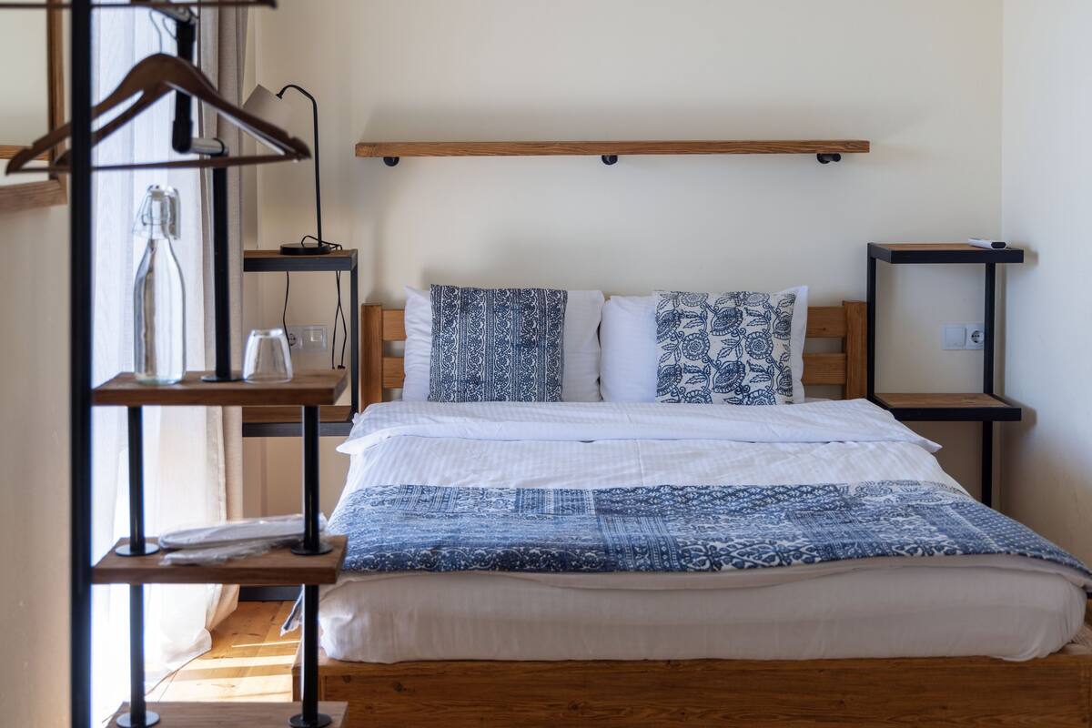 Un lit bien fait, recouvert d’une couvre-lit à motifs bleus et blancs, avec une tête de lit en bois et deux tables de chevet assorties, chacune dotée d’une petite étagère.
