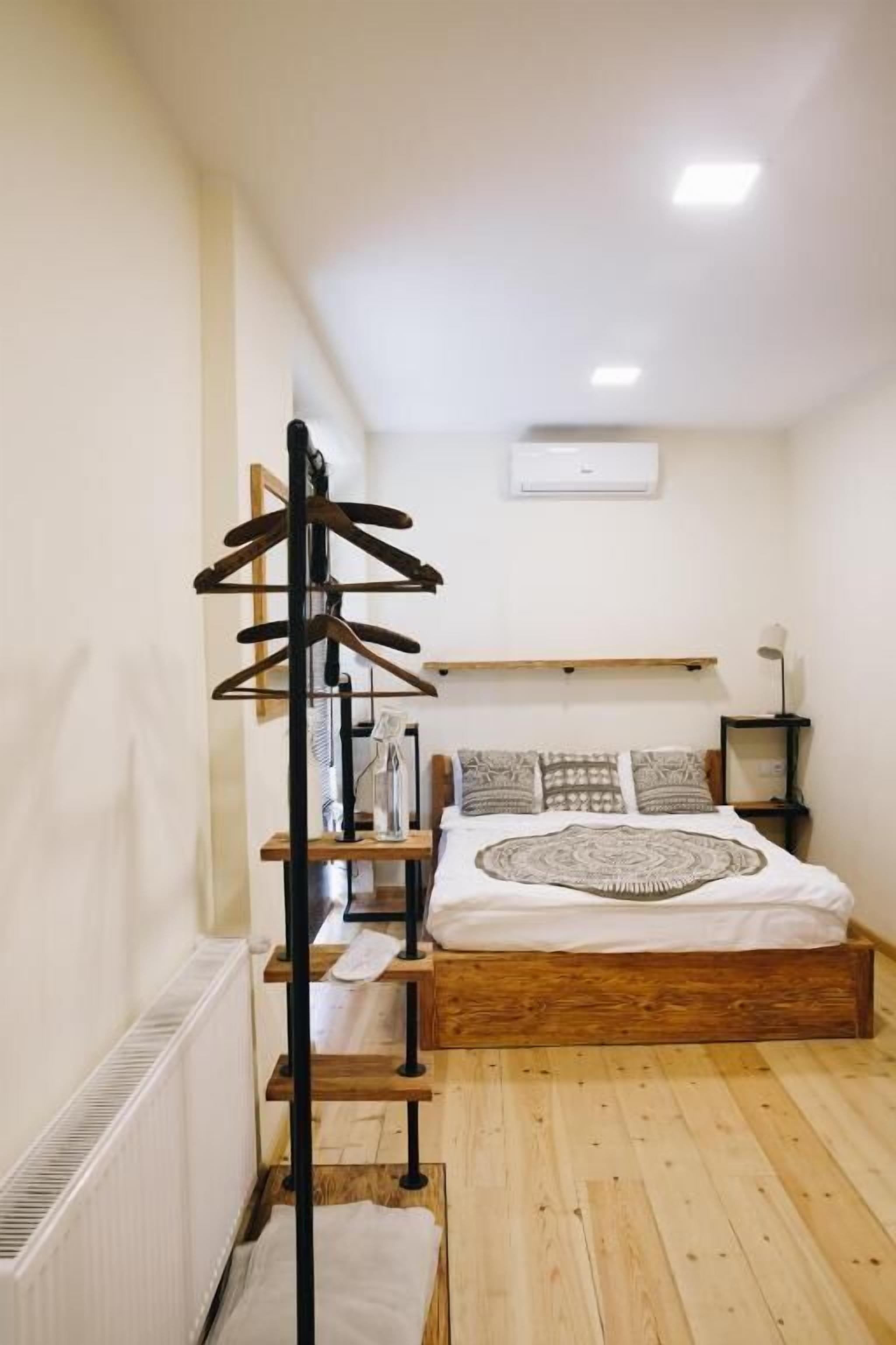 A bedroom with a wooden floor, a bed with patterned bedding, a wooden coat rack, and a small table with a lamp.