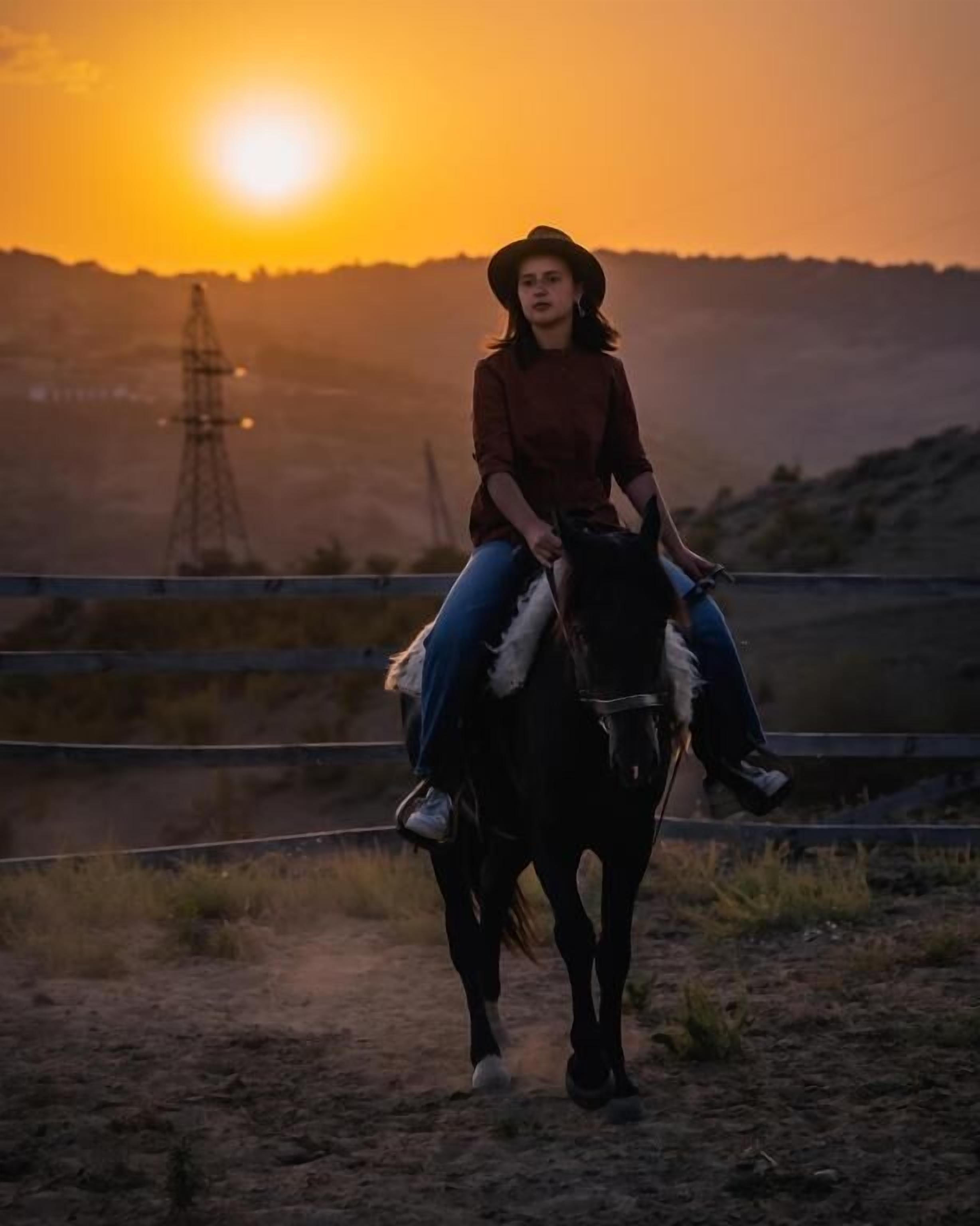 Eine Person reitet auf einem Pferd einen Feldweg entlang, im Hintergrund ein Sonnenuntergang.