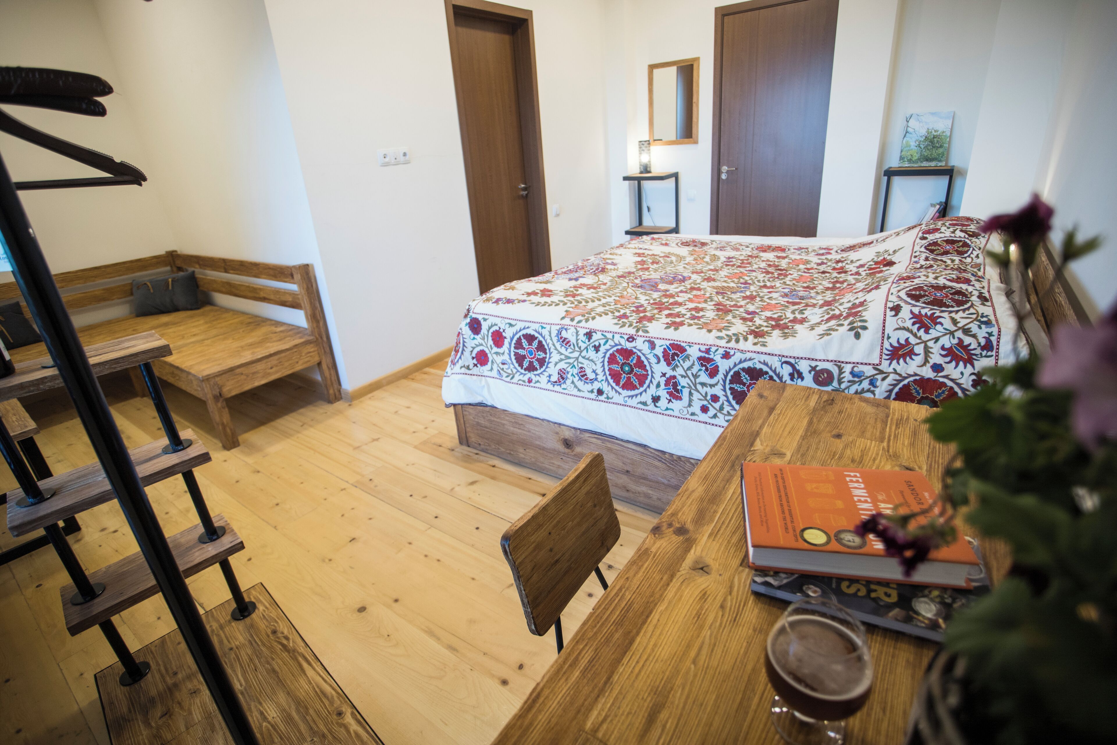 A bedroom with a wooden bed, a bedside table with a book and a glass, a chair, and a wooden ladder.