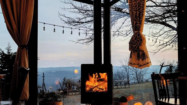 A cozy room with a wood-burning stove, wooden furniture, and a view of a landscape. - Lost Ridge Inn & Brewery & Ranch (Sighnaghi)