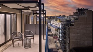 A balcony with two chairs and a small table, overlooking a cityscape with buildings and a waterfront. - Athenarum Portus Life & Style Hotel (Piraeus)