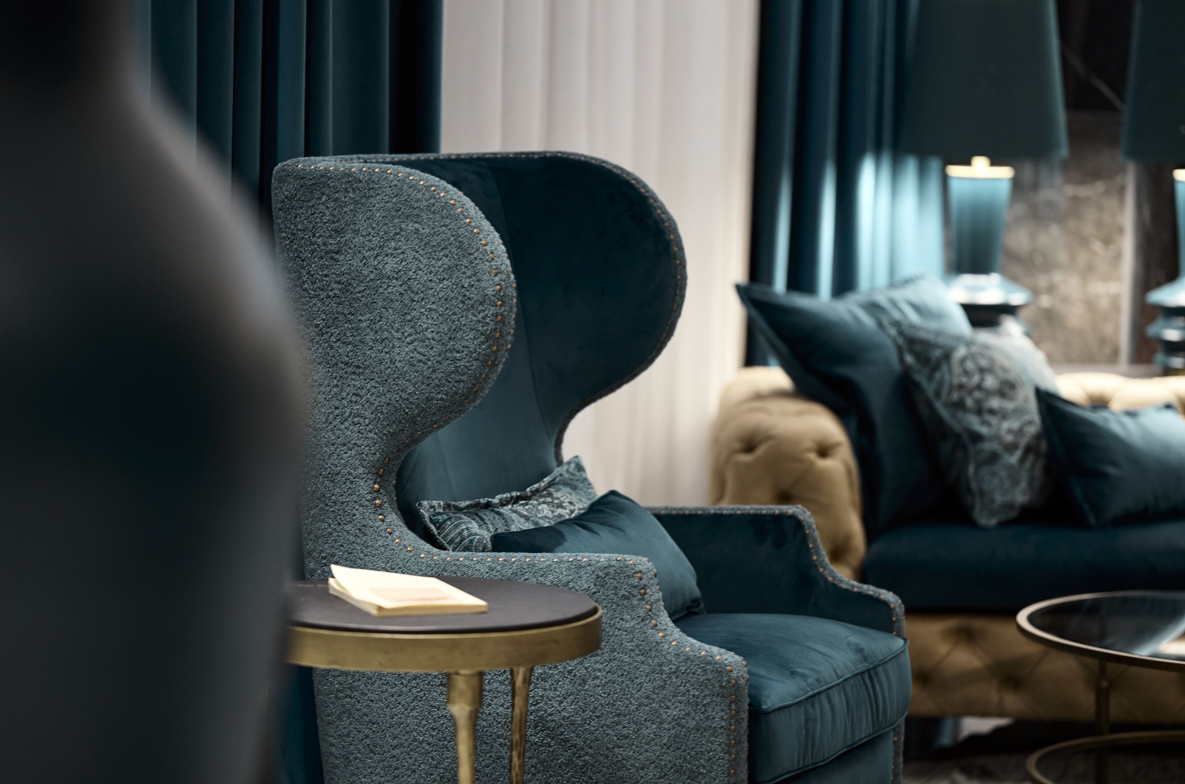 A modern living room with a dark blue armchair, a small round table, and a sofa with patterned cushions.