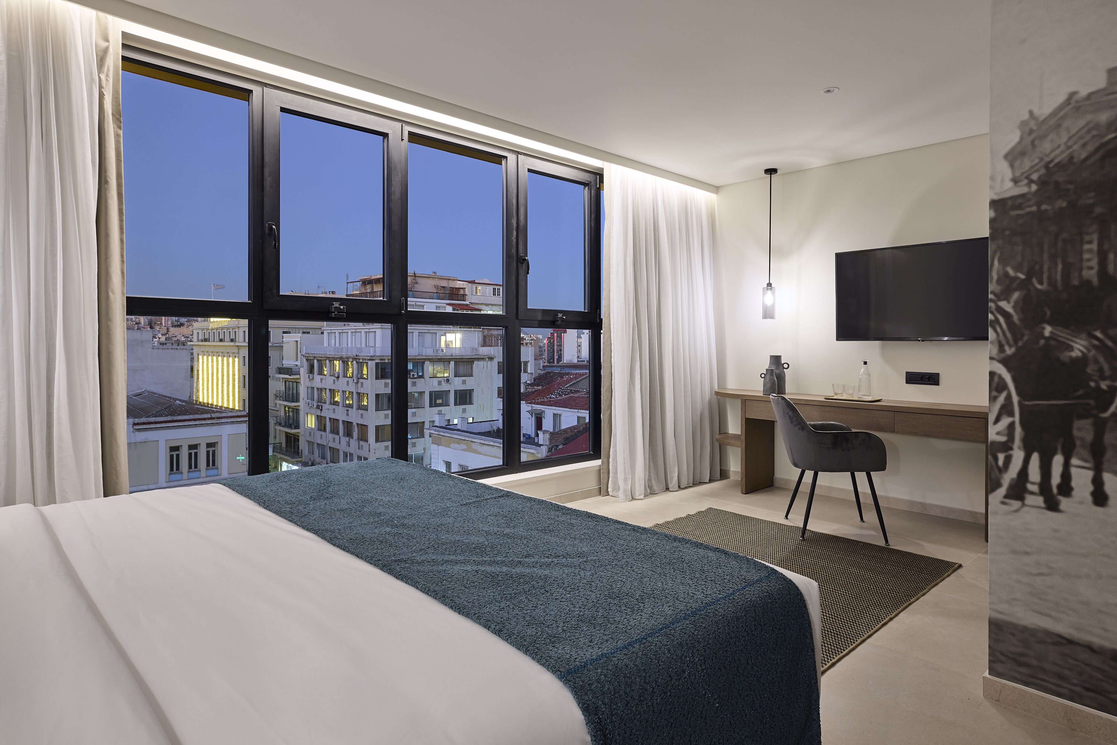 A hotel room with a large window, a bed, a desk, a chair, and a flat-screen TV.