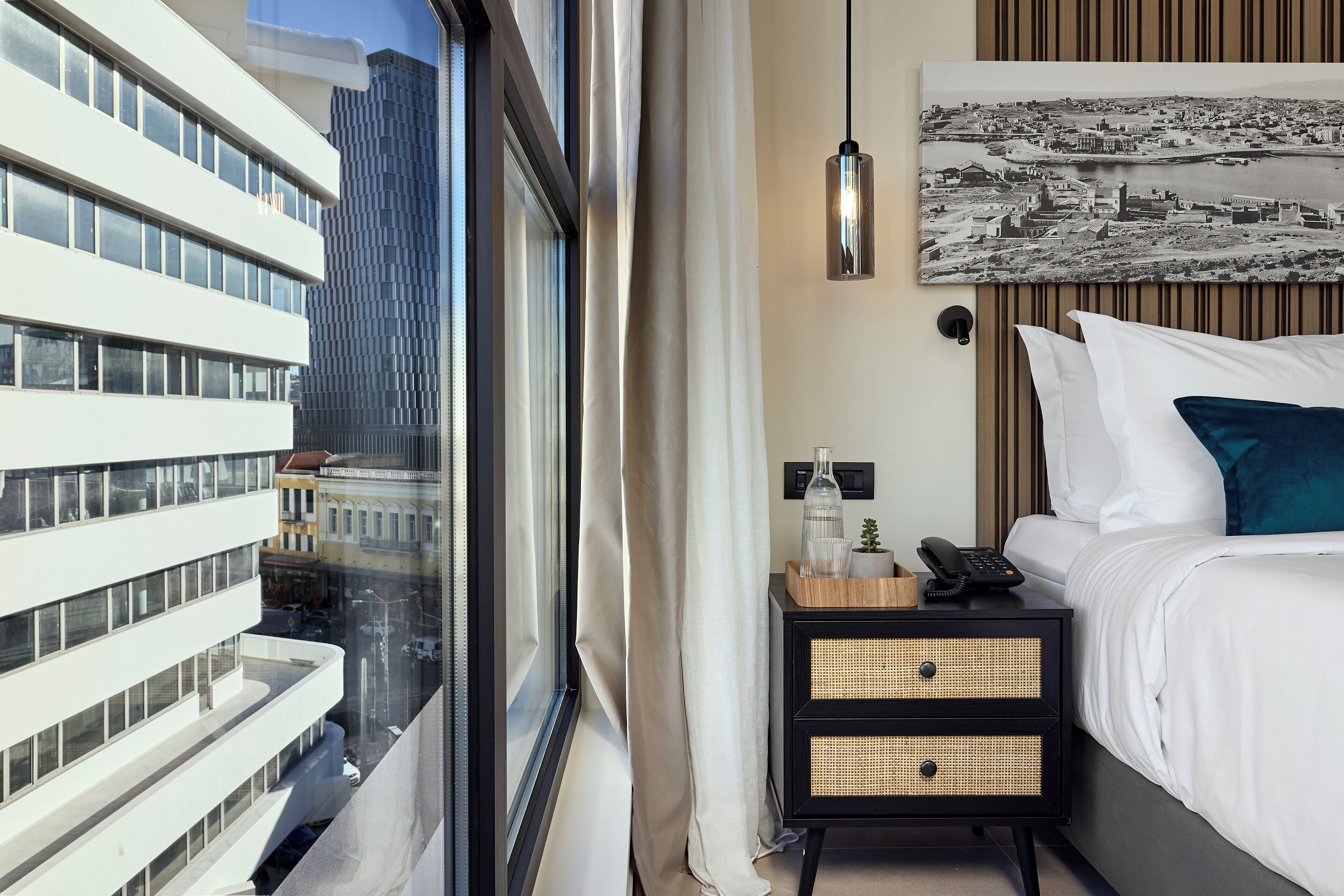 Hotelkamer met een groot raam, een bed met witte lakens, een nachtkastje met telefoon en uitzicht op de stad.