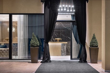 A hotel lobby with a marble reception desk, large windows with sheer curtains, and two potted plants.