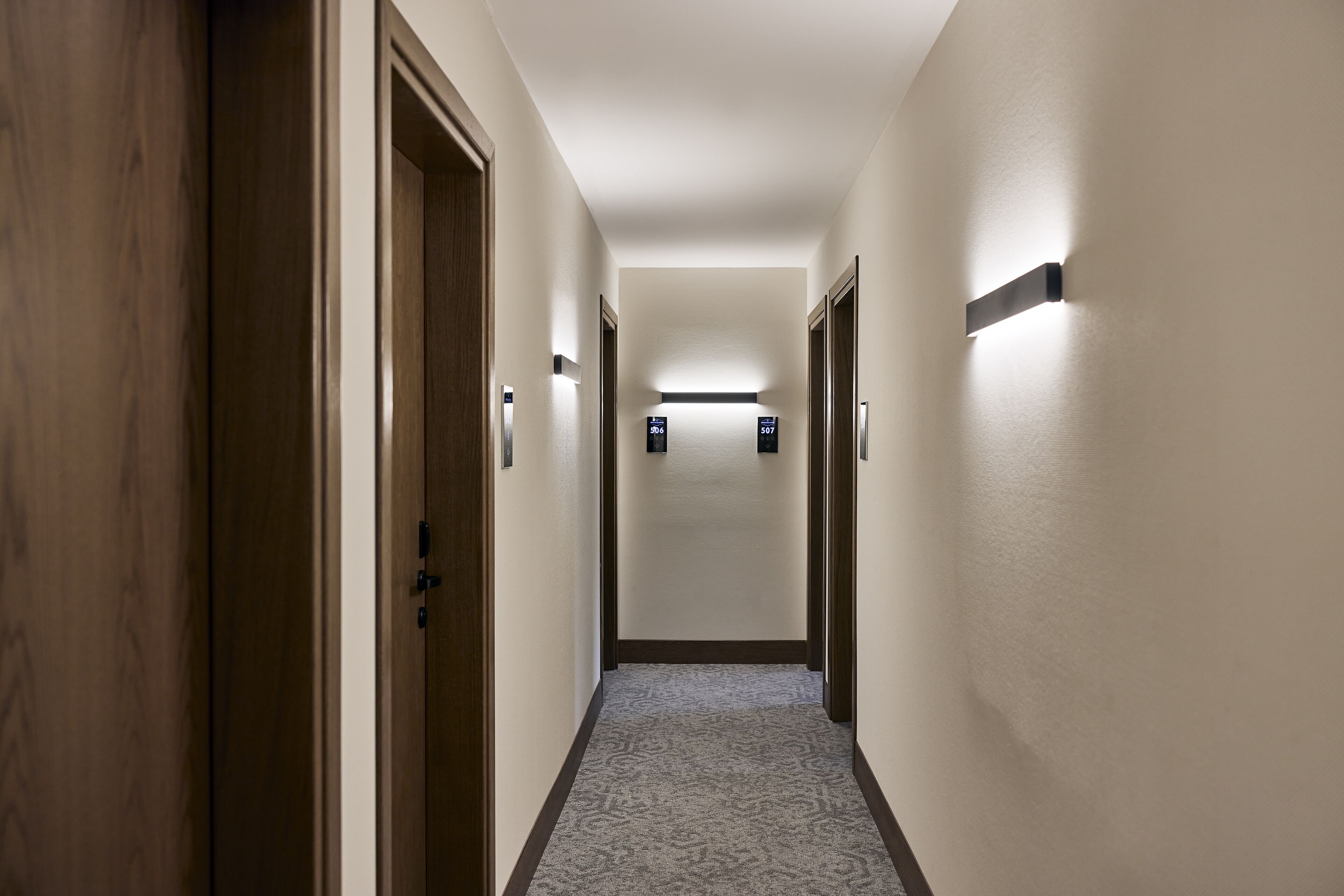 A narrow, carpeted hallway with wooden doors on one side and a wall-mounted light fixture on the other.