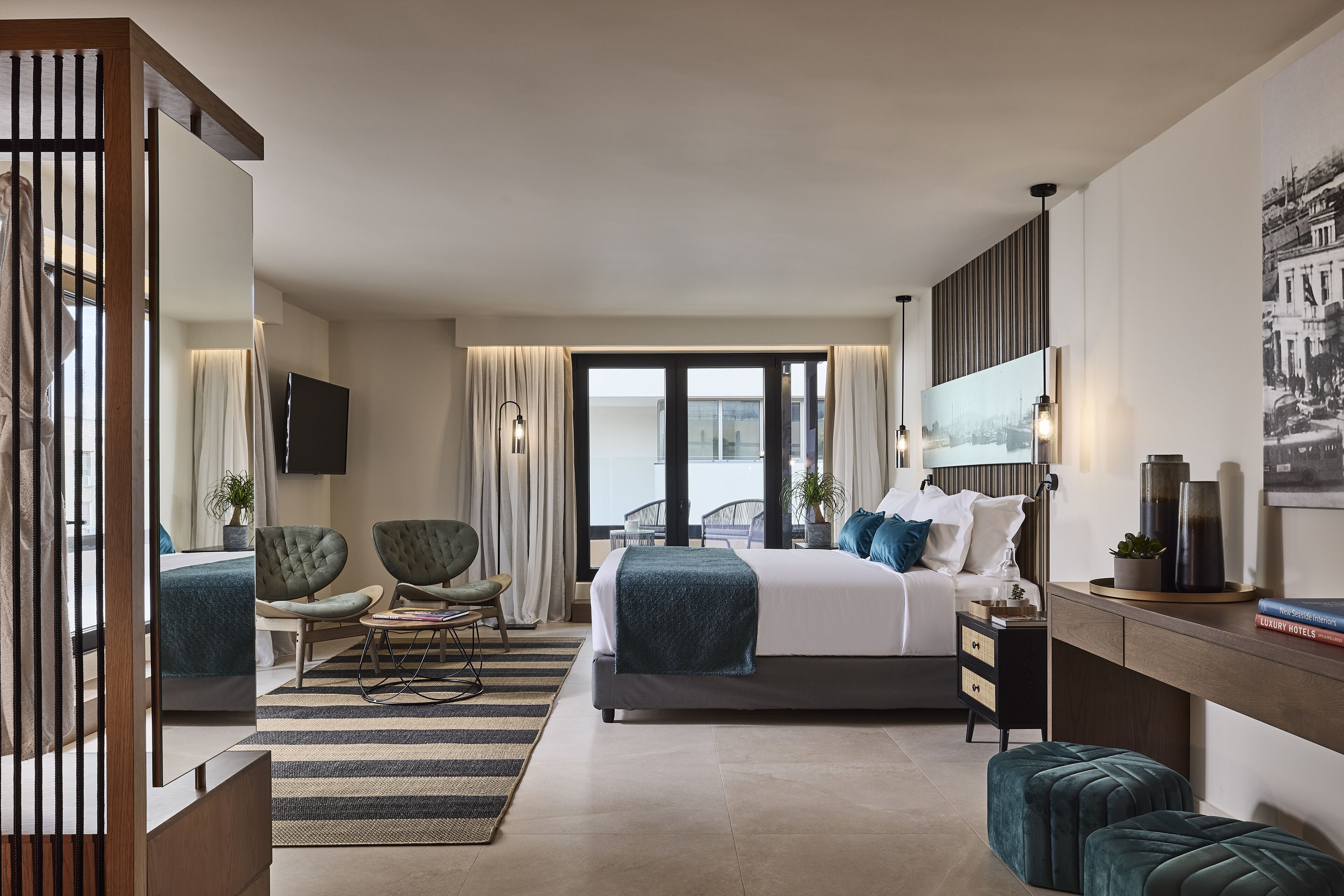 Camera d'albergo moderna con un letto grande, due poltrone, una scrivania e vista sulla città attraverso ampie finestre.