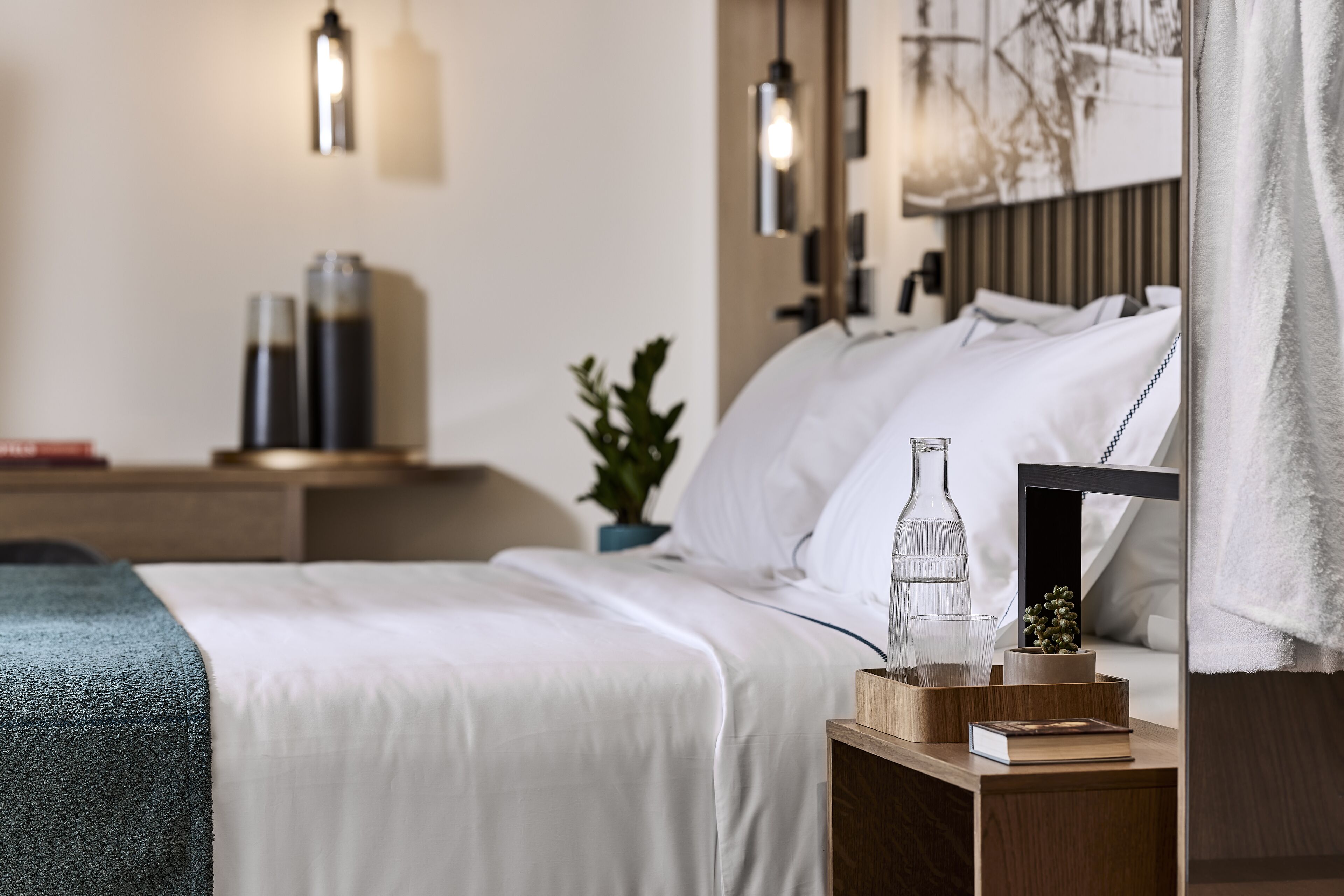 A neatly made bed with white linens, a bedside table with a book and a glass, and a wall-mounted lamp.