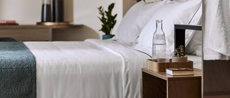 A neatly made bed with white linens, a bedside table with a book and a glass, and a wall-mounted lamp.