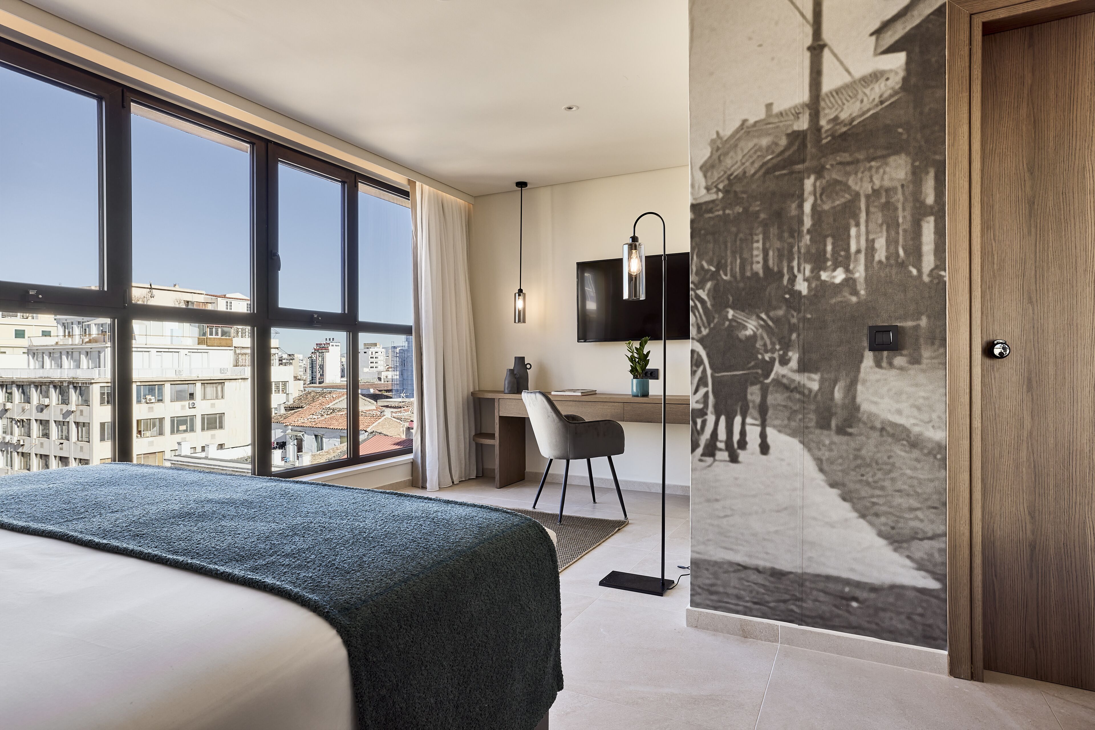 Habitación de hotel moderna con una cama grande, un escritorio, una silla, un televisor y vistas a la ciudad a través de grandes ventanales.