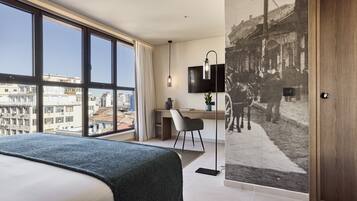 A modern hotel room with a large bed, a desk, a chair, a TV, and a city view through large windows.