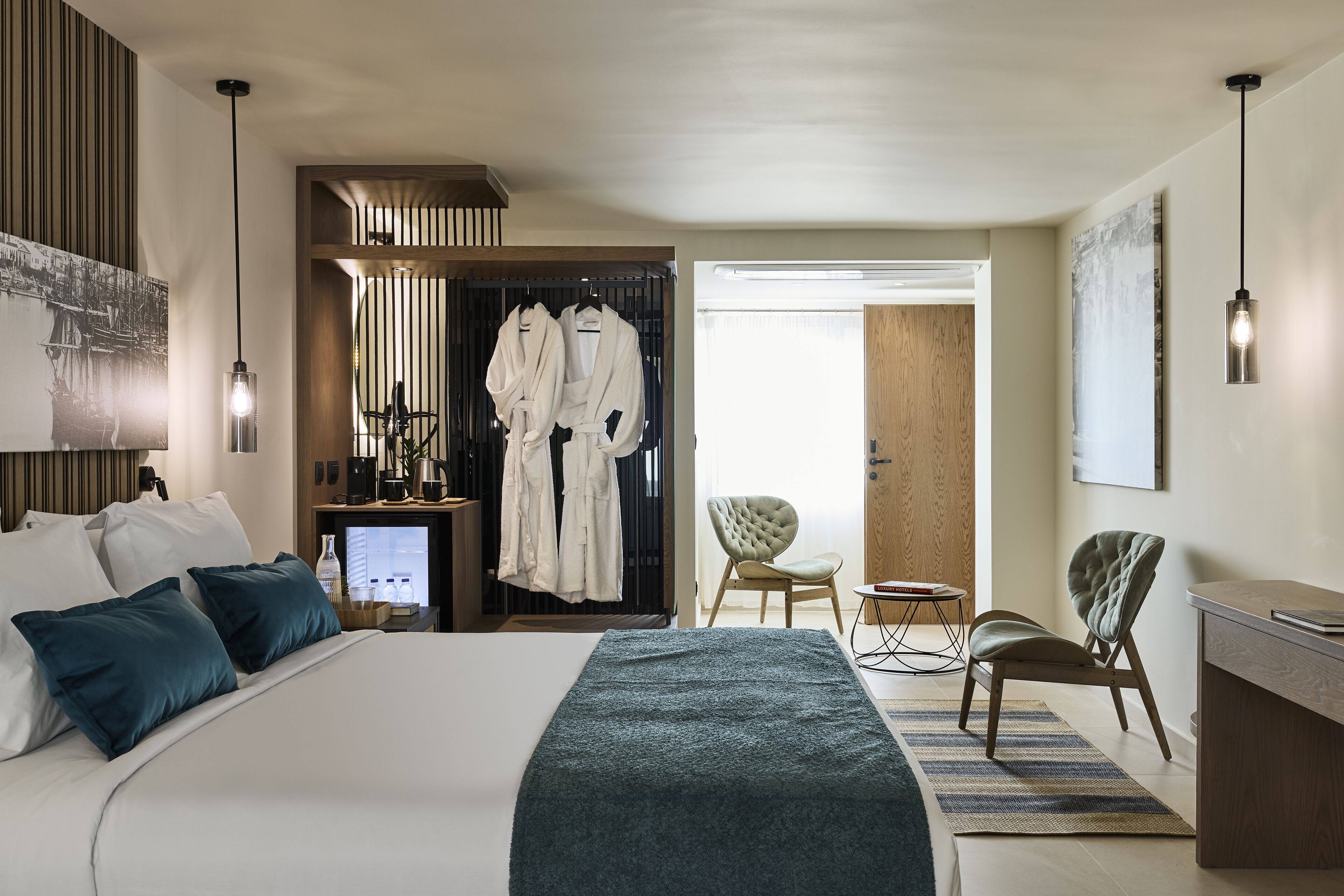 A modern hotel room with a large bed, a desk, two chairs, a robe rack, and a coffee station.