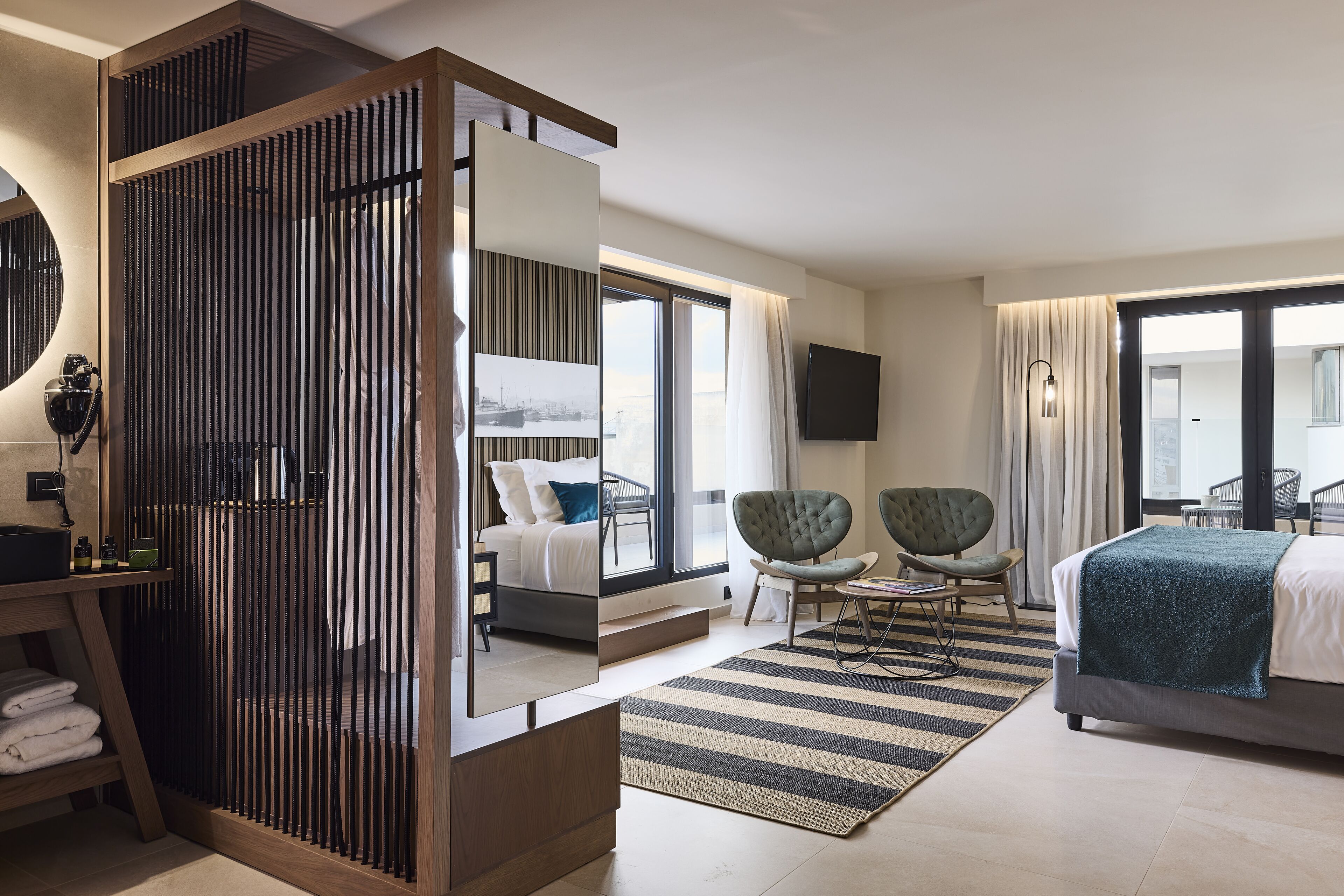 A modern hotel room with a large bed, a seating area with two chairs, a large mirror, and a view of the city through the window.