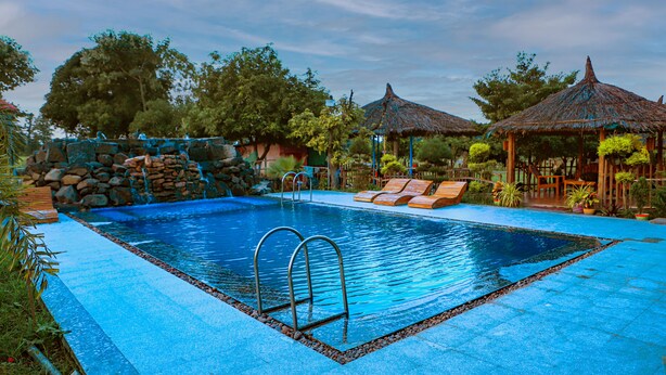 Una piscina rectangular con borde de piedra, rodeada de sillas de descanso y construcciones con techo de paja.