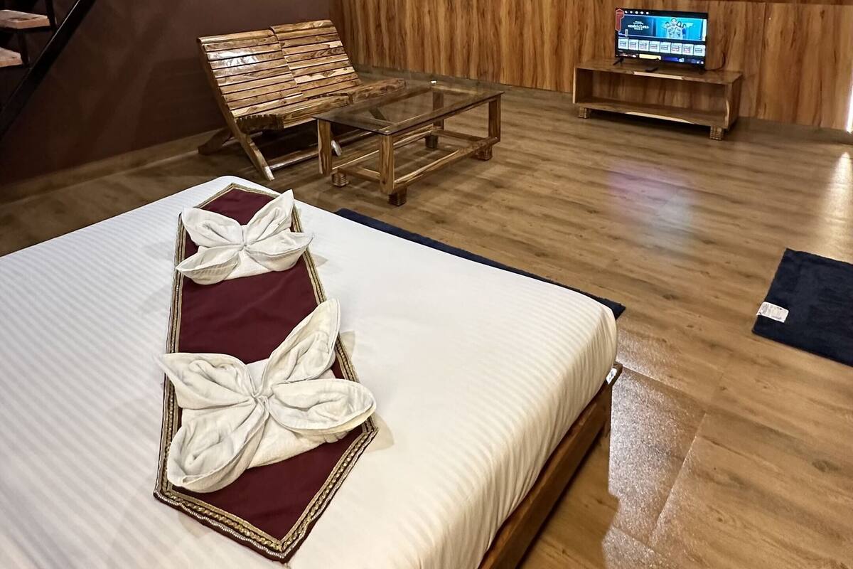 Un dormitorio con piso de madera, una cama con cabecera roja, un banco de madera, una mesa de centro de vidrio, un televisor sobre un mueble de madera y una escalera.