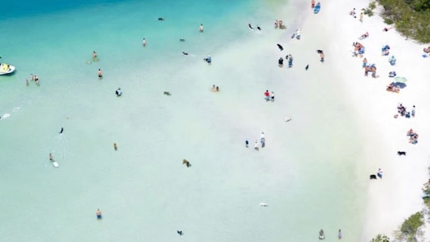 Luchtbeeld van een strand met helder turquoise water, een boot en mensen aan de kust.