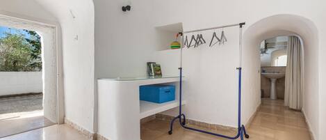 Quarto com escrivaninha branca, caixa de armazenamento azul, varal de roupas e vista para um banheiro.