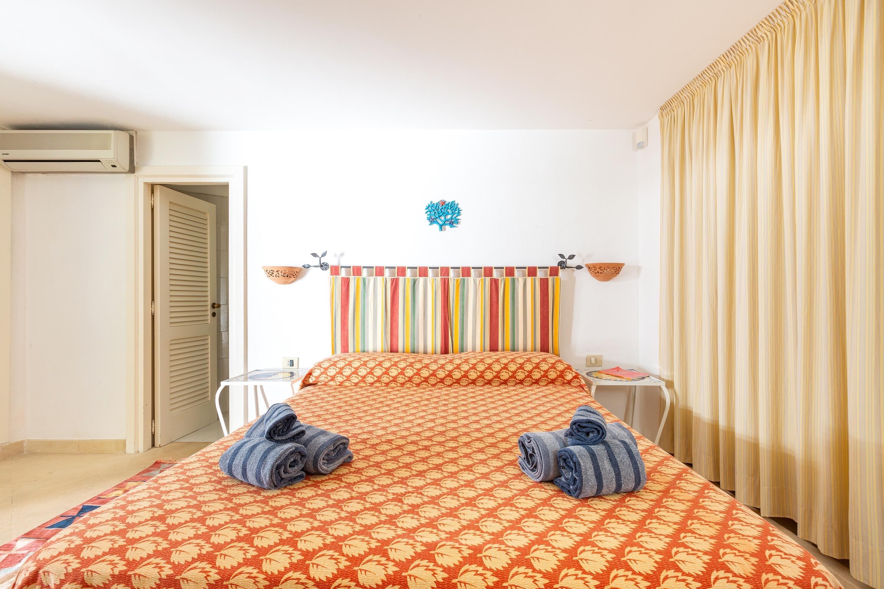 A hotel room with a bed, a small table, a chair, a wall-mounted air conditioner, and a curtain.