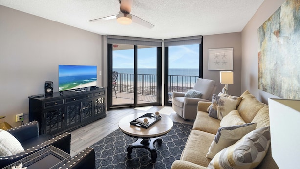 A modern living room with a large TV, a black entertainment unit, a round coffee table, a beige sofa, and a view of the beach through large sliding doors. - Phoenix VII 713 by Liquid Life (Orange Beach)