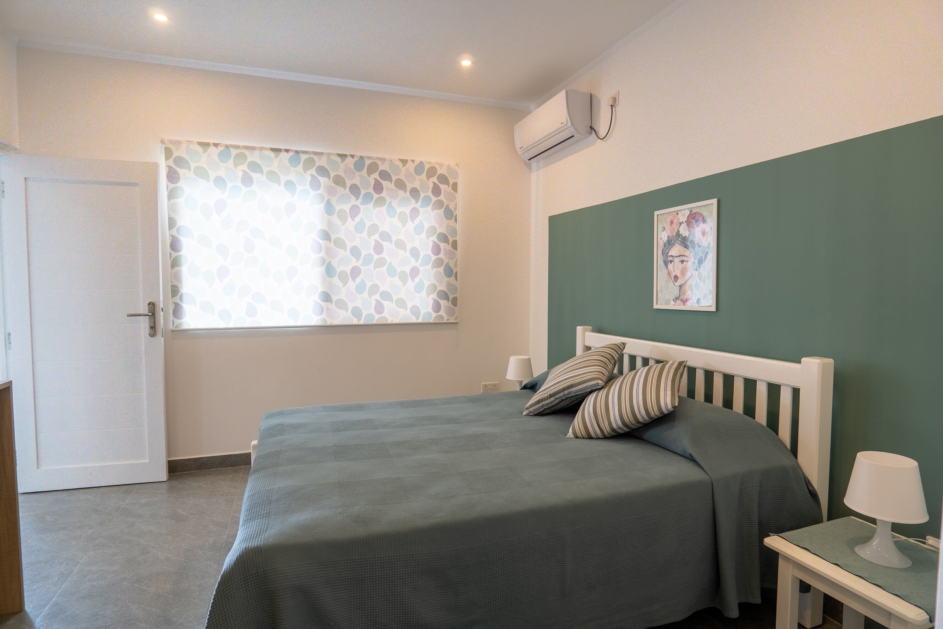 Un dormitorio con cama, mesita de noche con lámpara, ventana con cortinas estampadas y un equipo de aire acondicionado en la pared.