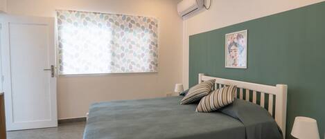 Un dormitorio con cama, mesita de noche con lámpara, ventana con cortinas estampadas y un equipo de aire acondicionado en la pared.