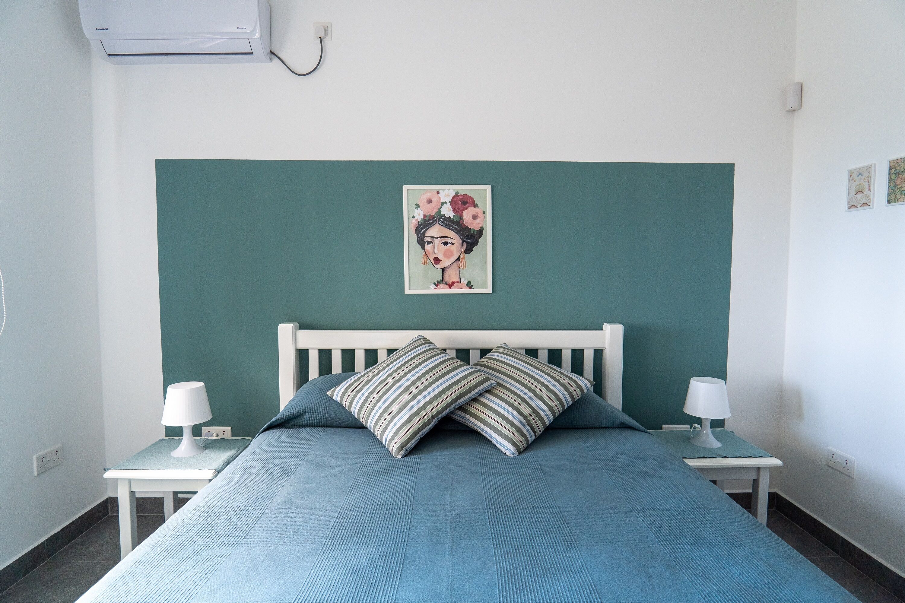 A bedroom with a teal headboard, a bed with blue bedding, two bedside tables with lamps, and a wall-mounted air conditioner.
