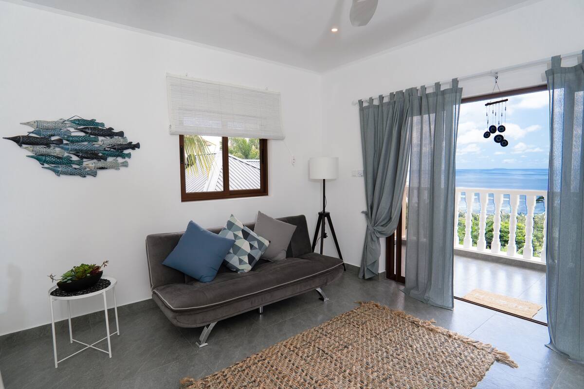 Ein Wohnzimmer mit grauem Sofa, einem kleinen weißen Tisch mit Pflanze, einem großen gewebten Teppich, einer Dreibeinlampe und Meerblick durch einen Balkon mit grauen Vorhängen.