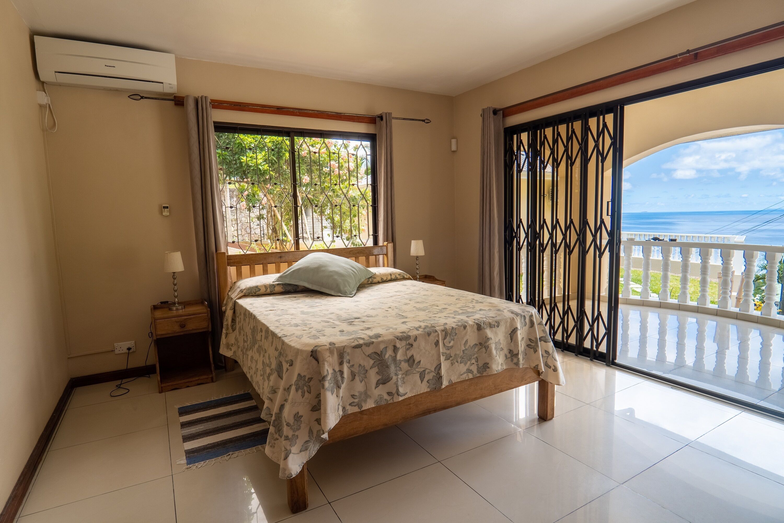 Ein Schlafzimmer mit Bett, Nachttisch, Fenster mit Vorhängen, Meerblick und einem Balkon mit Geländer.