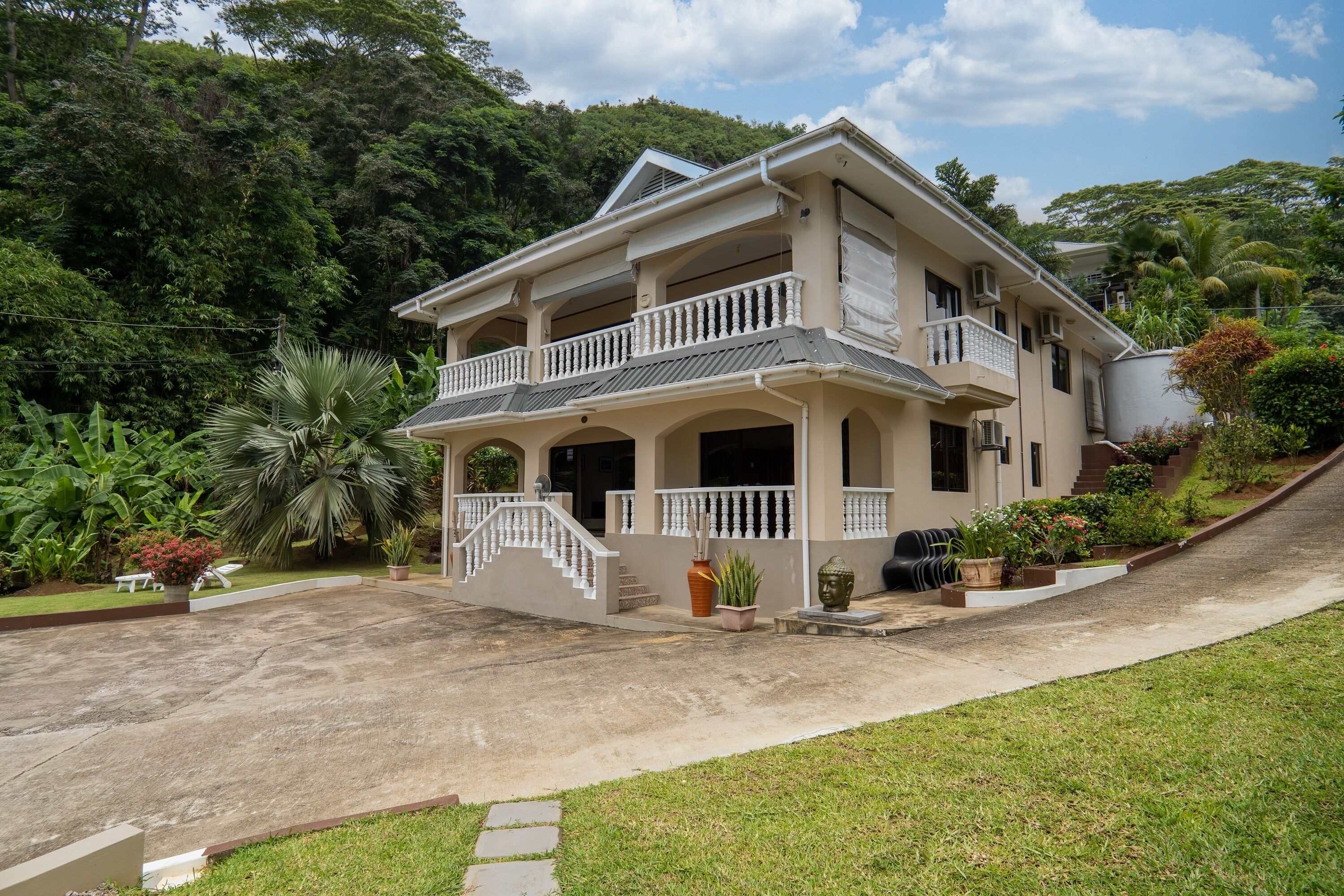 A two-story house with a balcony, surrounded by lush greenery and a paved driveway.