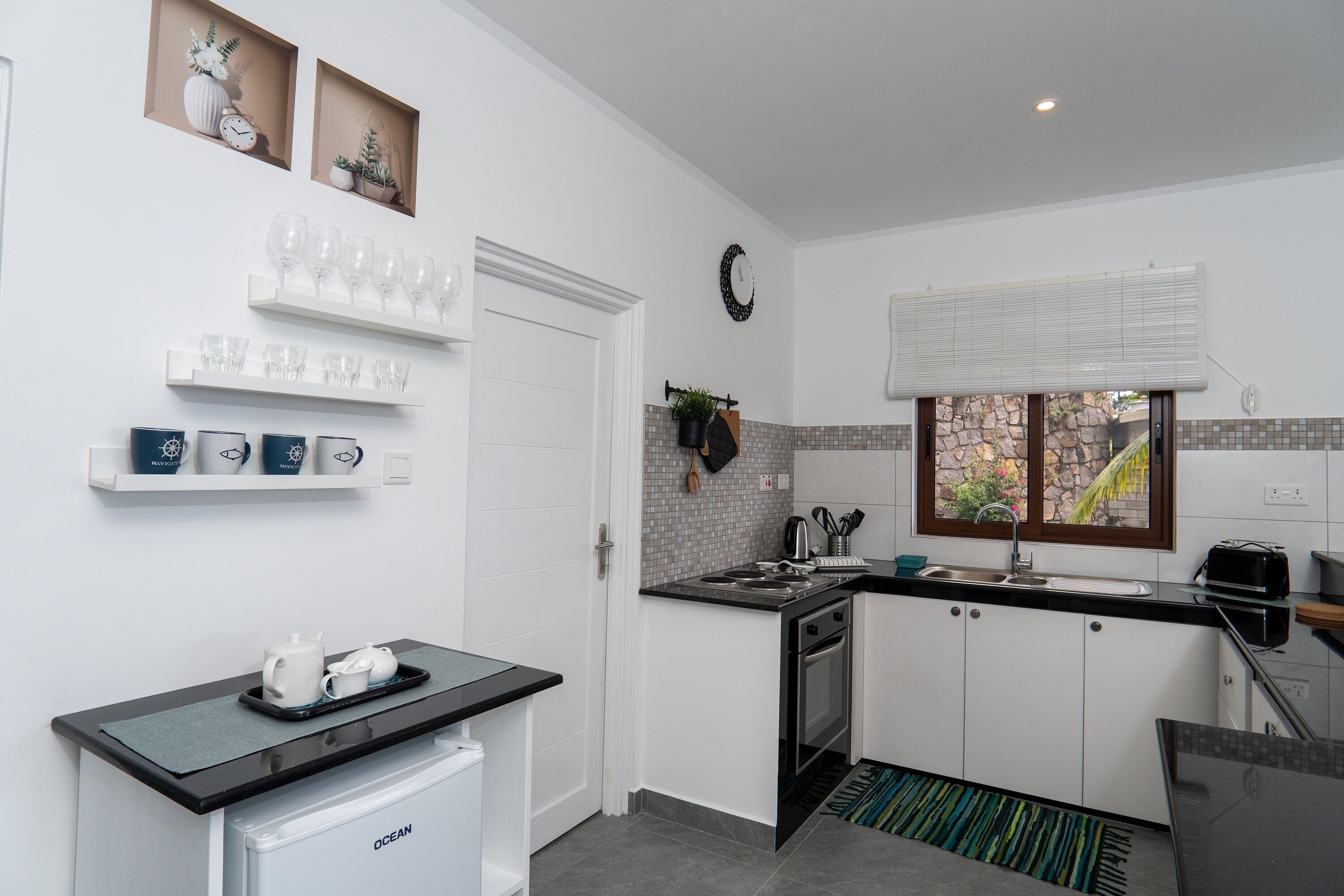 A modern kitchen with white cabinets, a built-in oven, and a small refrigerator. There are shelves with glassware and a window with a view of greenery.