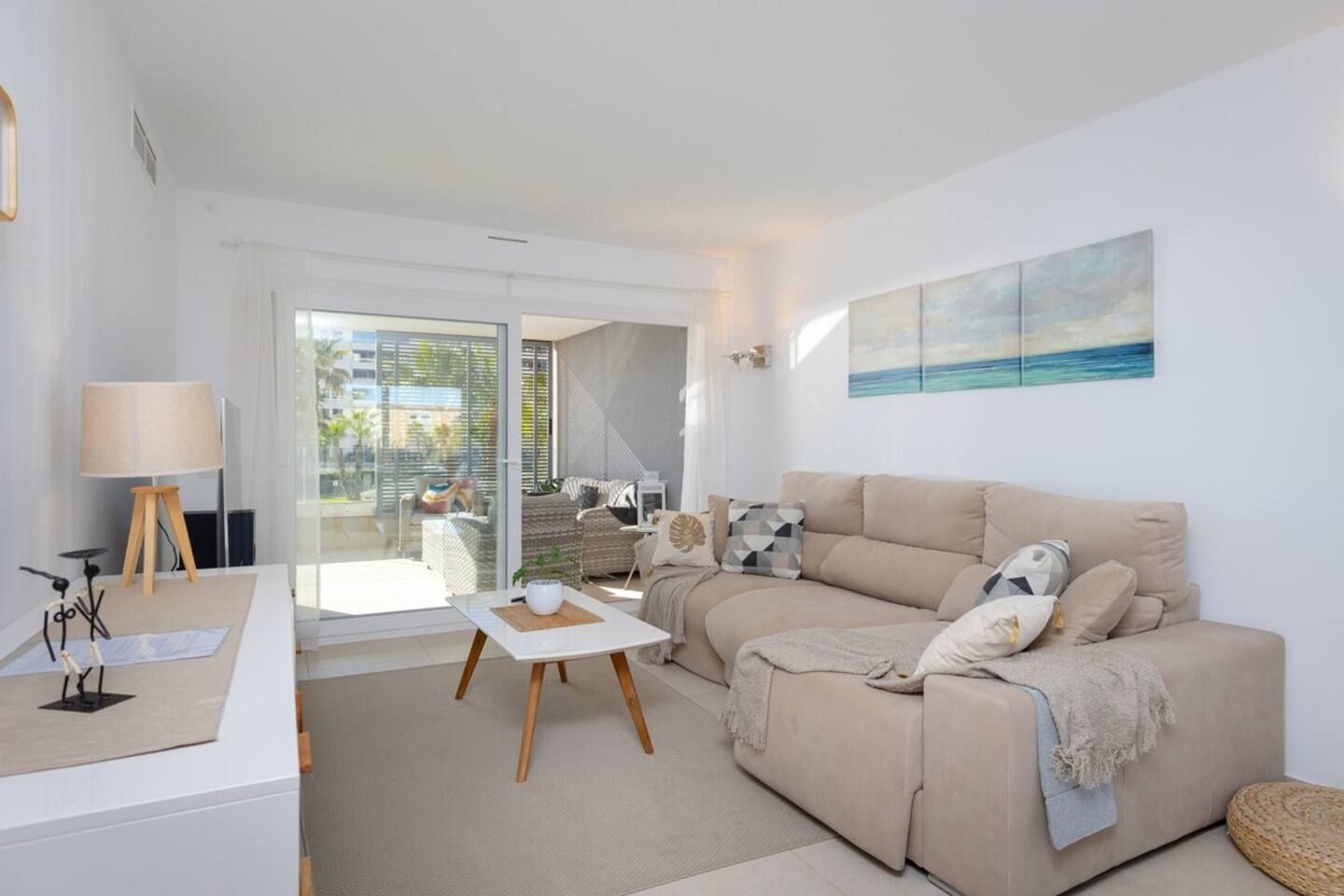 A modern living room with a sofa, a coffee table, and a lamp. There is a painting on the wall and a view of the outdoors through a glass door.