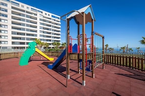 Ein Spielplatz mit Rutsche und Klettergerüst, gelegen in einem befestigten Bereich mit Blick auf ein Gebäude und das Meer.