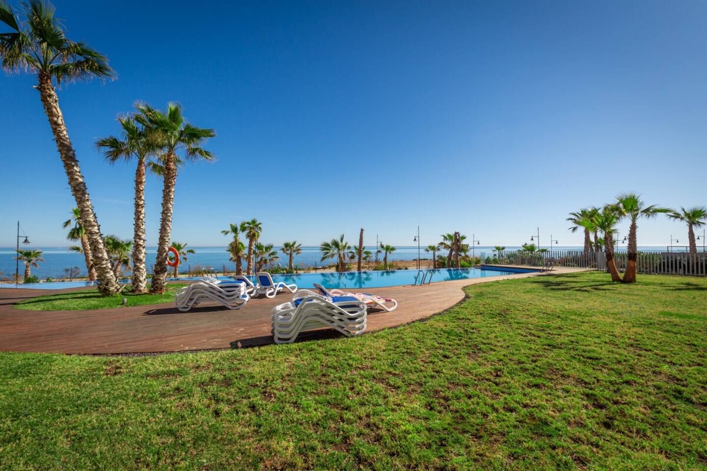 A pool area with lounge chairs and palm trees, overlooking a body of water.
