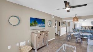 A spacious living room with a large TV, a dining area with a table and chairs, and a kitchen in the background. - Perdido Dunes 29 by Liquid Life (Orange Beach)
