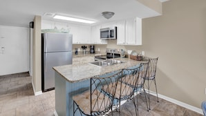 A modern kitchen with white cabinets, a stainless steel refrigerator, a microwave, and a granite countertop. There are two bar stools at the island. - Perdido Dunes 29 by Liquid Life (Orange Beach)